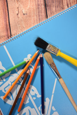 Paint brushes and pencils on a blue notebook on a wooden background
