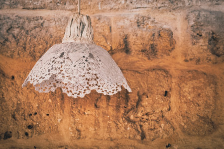 A vintage decorative lamp covered in lace standing in front of a tumbled stone wall background. Home...