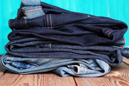 Stack of blue and dark blue jeans lying on a wooden table with bright turquoise backgroundの写真素材