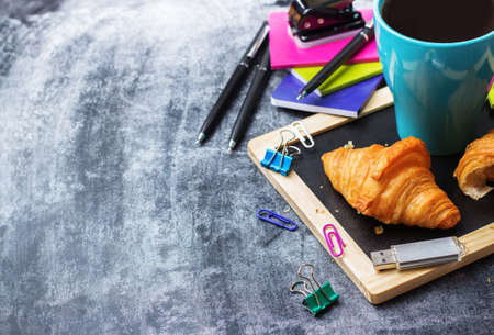 Education, back to school, business office breakfast concept. Stationery, supplies, pencil, pen, note, croissant and mug of black coffee on a grunge chalkboard. Selective focus, copy space backgroundの写真素材