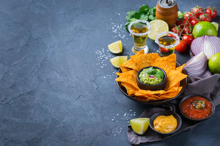 Yellow corn tortilla chips nachos with guacamole, red hot jalapeno chili salsa and cheese sauce with tequila on a dark table. Copy space background の写真素材