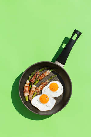 Balanced diet nutrition keto food concept. Healthy ketogenic low carb fried eggs, bacon, asparagus in a cooking pan. Trendy sunny, vivid, harsh and hard light and shadows on a green mint backgroundの写真素材
