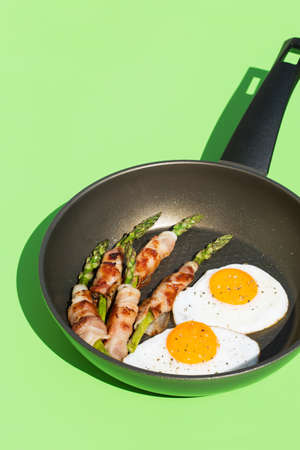 Balanced diet nutrition keto food concept. Healthy ketogenic low carb fried eggs, bacon, asparagus in a cooking pan. Trendy sunny, vivid, harsh and hard light and shadows on a green mint backgroundの写真素材