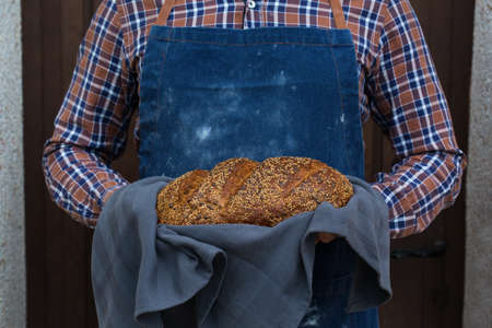 Young man baker holding freshly baked loaf of homemade rye bread in his hands