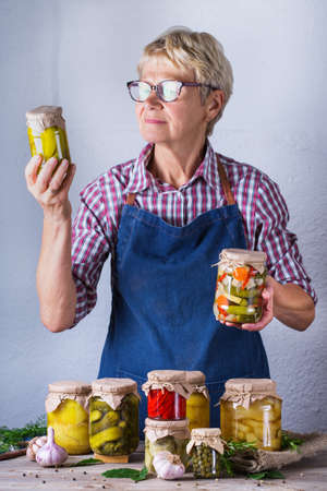 Senior mature woman holding in hands jar with homemade preserved and fermented food. Variety of pickled and marinated vegetables. Housekeeping, home economics, harvest preservation  の写真素材