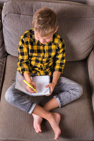 Cute smiling boy sitting on a couch and writing in his notebook. Stay at home, lockdown, social distancing, quarantine.の写真素材