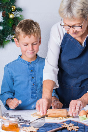 Happy senior mature woman, grandmother and young boy, grandson cooking, kneading dough, baking pie, cake,  cookies. Family time in the cozy kitchen. Seasonal winter Christmas activity at home.の写真素材