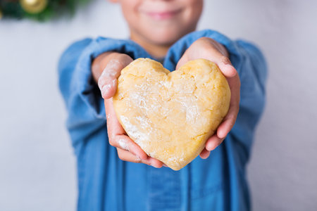 Happy cute young boy holding in hands heart shaped piece of homemade dough. Family time in the cozy kitchen. Winter activity at home. Help and homework training for kidsの写真素材