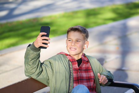 Handsome teenager waving hand saying hello sitting on a bench on the city street, outdoors. Caucasian young boy is talking with his friends on the phoneの写真素材