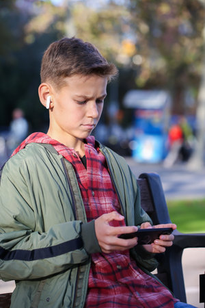 Handsome teenager looking to his phone sitting on a bench on the city street, outdoors. Caucasian young boy is waiting his friends for a walkの写真素材