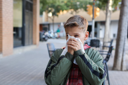 Sick and cold teenager blowing nose and sneezing sitting in outdoor cafe. Caucasian young boy is suffering from fluの写真素材