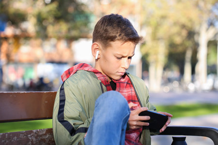 Handsome teenager looking to his phone sitting on a bench on the city street, outdoors. Caucasian young boy is waiting his friends for a walkの写真素材