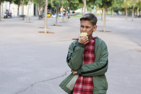 Handsome teenager eating green apple on the city street, outdoors. Caucasian young boy is waiting his friends for a walkの写真素材