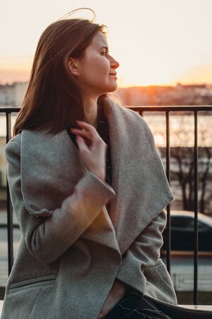 Caucasian brown-haired girl in a beige coat straightens hair in the wind. sunset behindの写真素材