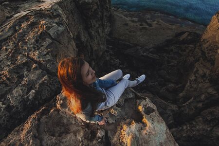 Woman traveler sitting near sea on top of cliff in summer mountains and enjoying view of sea and nature. Cape Greco, Cyprus, Mediterranean Sea. Sunriseの写真素材