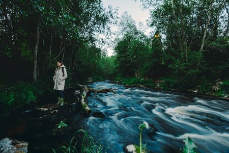 Young woman staying in the middle of the stream of a river near fallen broken tree in the summer forestの写真素材