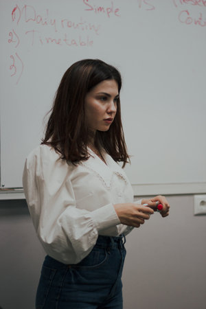 Young female english language teacher with a red marker in a white blouse standing in front of a...