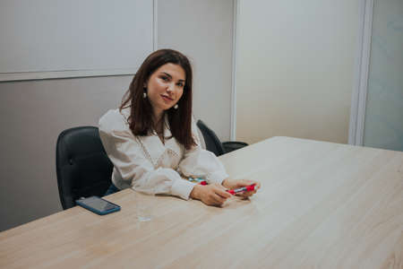 Young brunette woman in a white blouse sitting at the table in the office and holding a marker in...
