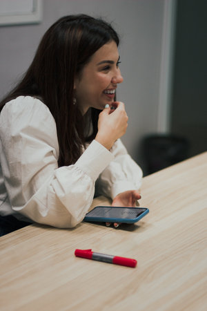 Young brunette woman in a white blouse sitting at the table in the office, holding a smartphone in...