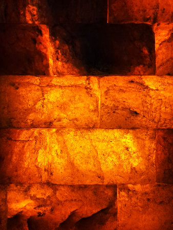 A striking close-up of a wall constructed from rough-cut salt blocks, illuminated from within to cast a rich, fiery orange and deep amber light. This texture creates a warm, atmospの写真素材