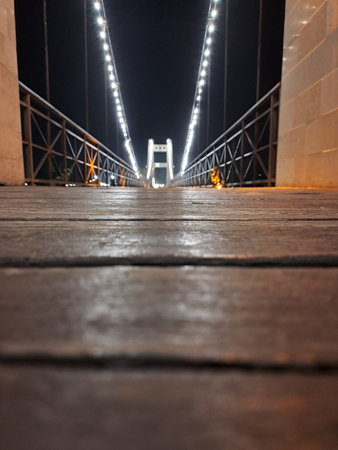An extremely low perspective shot emphasizing the rustic wooden planks of a footbridge extending towards a brightly lit central structure at nightの写真素材