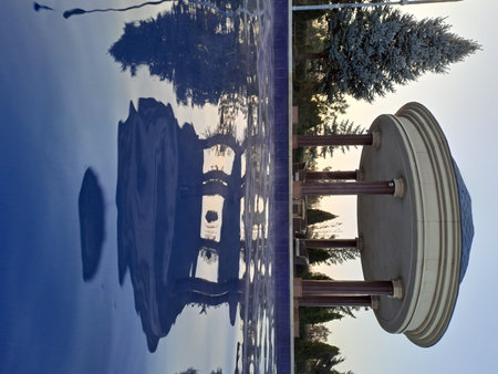 A tranquil scene showcasing a round open-air pavilion with columns and seating, perfectly reflected in the still, blue surface of a pool or pond under a clear sky at sunsetの写真素材