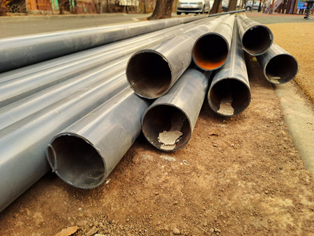 A close-up of several large, gray metal pipes lying on a dirt and concrete roadside, ready for...