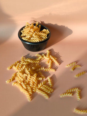 Dry fusilli pasta scattered on a peach colored surface next to a black bowlの写真素材