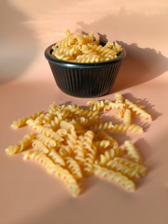 Dry fusilli pasta scattered on a peach colored surface next to a black bowlの写真素材