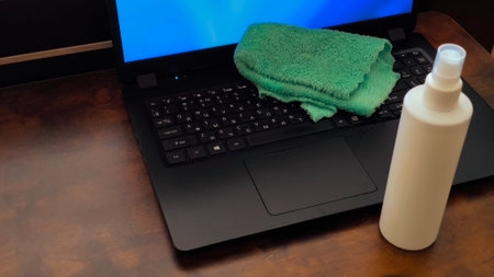 Laptop with cleaning cloth and spray bottle on a wooden table.