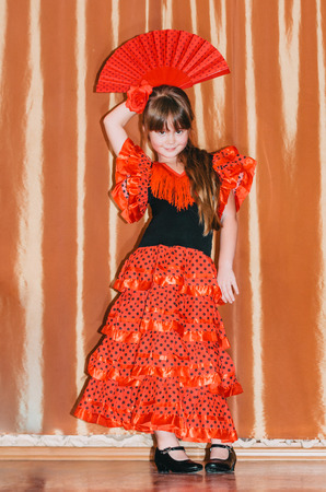 Cute little girl wearing beautiful red and black dress with matching headband, using hand fan, studio backgroundの写真素材