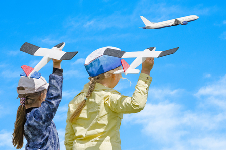 Two girls kid playing with toy airplane and a real passenger plane taking off in the sky, against blue summer sky background. Concept - inoculating the love of children for aviationの写真素材