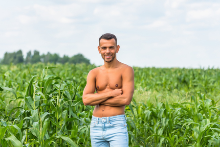 Young dark-skinned man smiles in light blue jeans in good physical shape stands in a rural spot in the green field of cornの写真素材