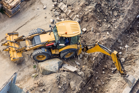 Yellow tractor with a bucket and back and front, digging a trench at a construction site. View from...