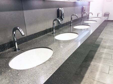 A row of washstands with a water tap and a soap dispenser in a public toilet, gray interior