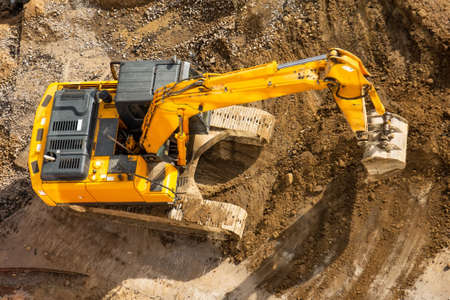 Excavator on the ground of a construction site with a raised bucket, top aerial view