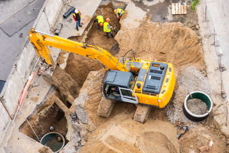 Excavator at a construction site while digging trenches for calcining sewer and drainpipes with a raised bucket, top aerial viewの写真素材