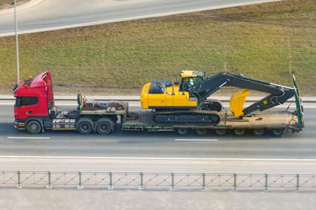 Heavy yellow excavator on transportation truck with long trailer platform on the highway in the city