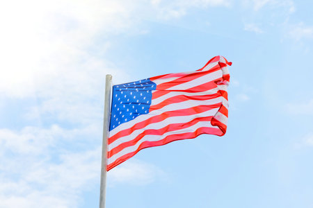 Tattered American flag blowing in the wind against a beautiful cloudscape and bright sunlightの写真素材