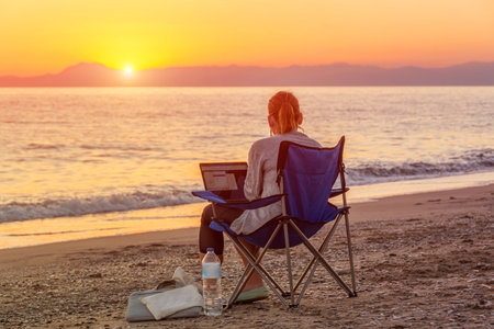 Girl with a laptop works online remotely on the coast of a sandy beach during a beautiful sunsetの写真素材