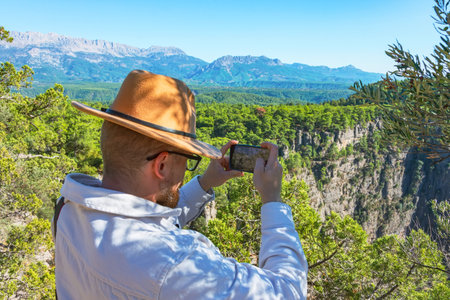 Caucasian male white shirt and brown hat takes pictures on smartphone of views on cliff near front of large canyon around the forest beautiful amazing view.の写真素材