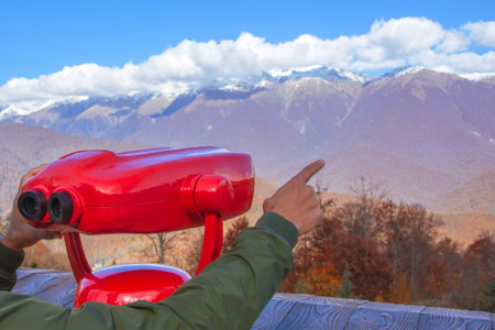 Young guy man points his finger at the mountain snow-capped peaks holding red binoculars for review and observation of panoramic nature viewsの写真素材