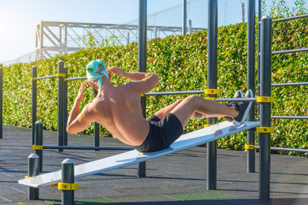 Muscular man in a bandana, bends the body and abs abdomen in different directions in an inclination on a bench in the park outdoorsの写真素材
