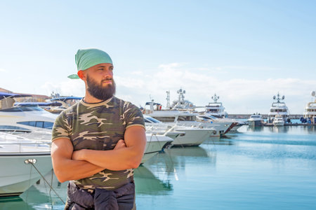 Handsome male beard turquoise bandanna military shirts stands hands folded on pier against the backdrop yachts in port bay in sunny weatherの写真素材