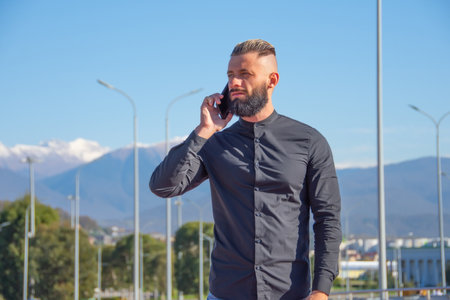 Man with a beard and slicked back hair in black shirt talking on phone, stands on the street of the...