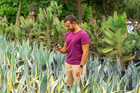 Young male gardener using smartphones takes orders and takes photos of plants to process customer orders in a garden among agaves and cacti. Sale of ornamental plants in the garden center, plantationの写真素材