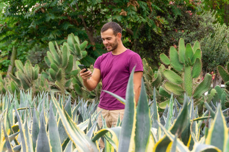 Young male gardener using smartphones takes orders and takes photos of plants to process customer orders in a garden among agaves and cacti. Sale of ornamental plants in the garden center, plantationの写真素材