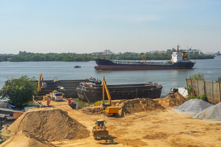 Excavator is loaded into a river vessel barges sand soil transportation of construction materials and materials along the river. Oil tanker floats nearby on the river.の写真素材