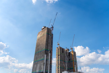 Couple standing high-rise towers in the scaffolding are covered with technical fabric with a protective tarpaulin with cranes on top, against the background of a blue sky with clouds.の写真素材