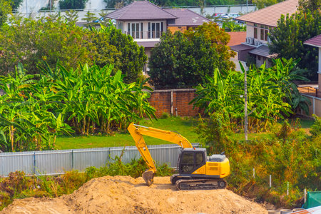 Excavator at work on construction site, banana thickets and other private houses around.の写真素材